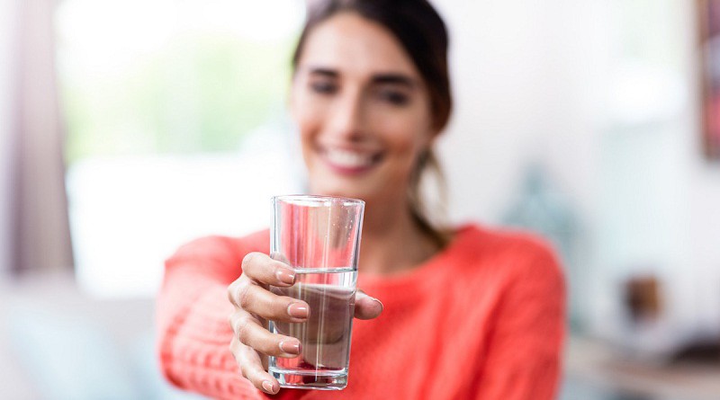 Happy young woman showing drinking glass with water at home